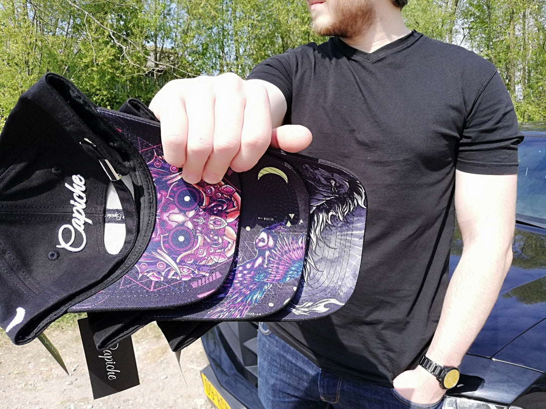 Close-up of a person displaying several Capiche men's caps with striking, colorful artistic designs on the brims. Premium headwear for men, showcasing detailed graphic artwork.