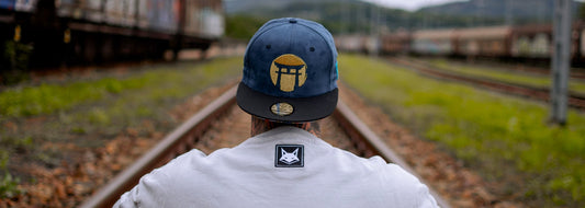 Une personne portant une casquette bleue avec un logo brodé en or, vue de dos, debout sur des voies ferrées, avec des bâtiments et de la verdure flous en arrière-plan.
