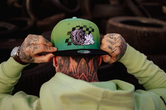 A Hip Hop hat from Capiche Caps in green ombre design featuring a smoking tire graphic, worn by a tattooed person adjusting the cap against a backdrop of stacked tires.