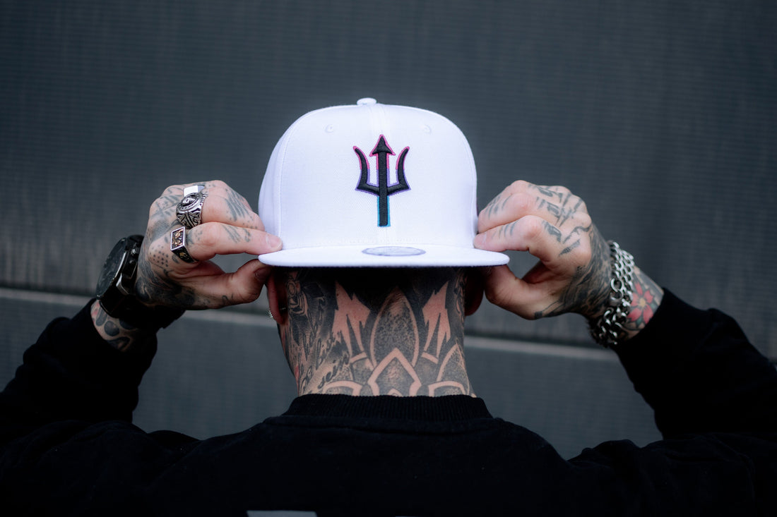 Close-up of a snapback white hat with a stylized purple and teal trident emblem, worn by an individual seen from behind.