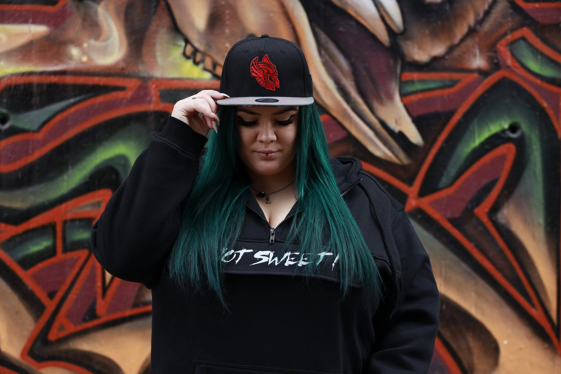 A woman with long teal-green hair wearing a black hoodie and a black snapback cap with a red embroidered design stands in front of a colorful graffiti wall, looking down while touching the brim of her cap.