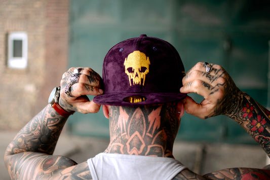 Un hombre sostiene una elegante gorra snapback de ante morado con un diseño de calavera dorada, mostrando una de las gorras con calavera únicas de Capiche Caps para hombre, perfecta para un estilo urbano atrevido.
