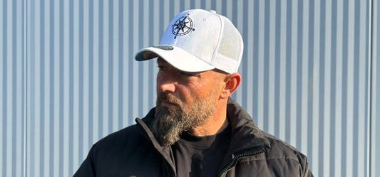Bearded man wearing a white mesh-back Capiche Caps hats for men trucker style cap with an embroidered front panel and curved brim, paired with a black jacket against a corrugated metal backdrop.