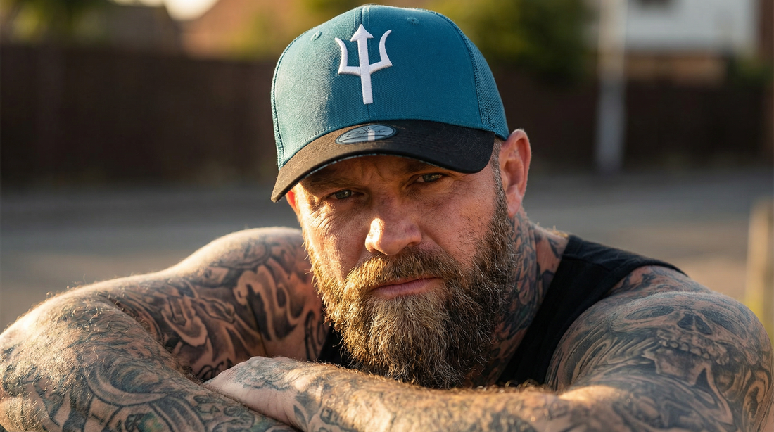 High quality trucker hats by Capiche Caps featuring a teal and black design with a bold trident logo, worn by a tattooed man outdoors in warm natural lighting.