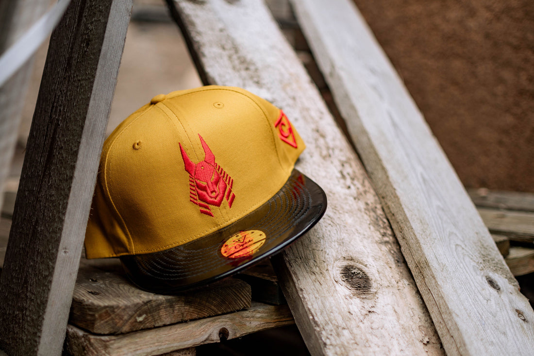 Yellow flat brim hat with a red embroidered Anubis logo and a black metallic brim, displayed on rustic wooden planks.