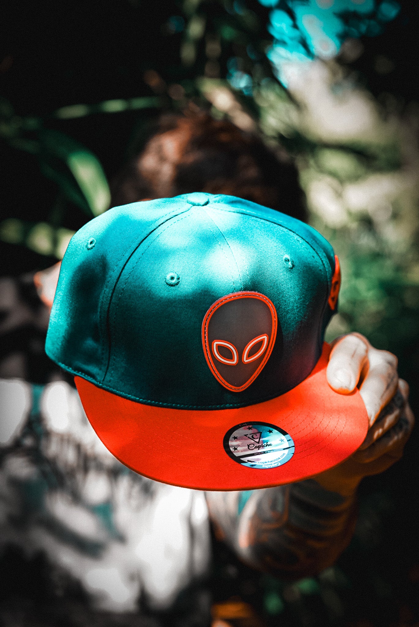 Tattoo Model Holding Blue Snapback Hat with Red Alien Front Logo From Capiche