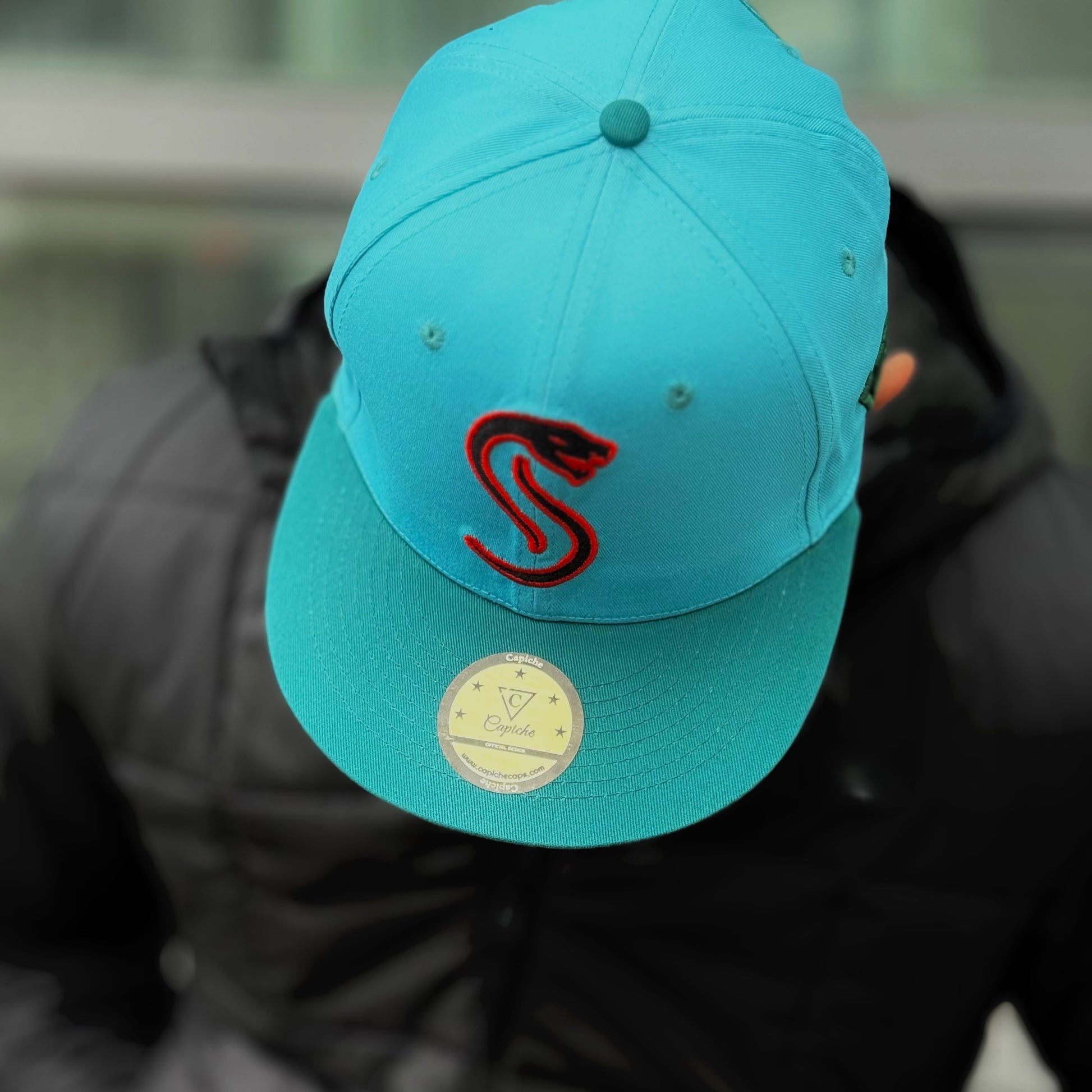 Man standing outside and showing the camera a close up of the blue snapback from Capiche with a red snake logo at the front and a golden sticker at the visor