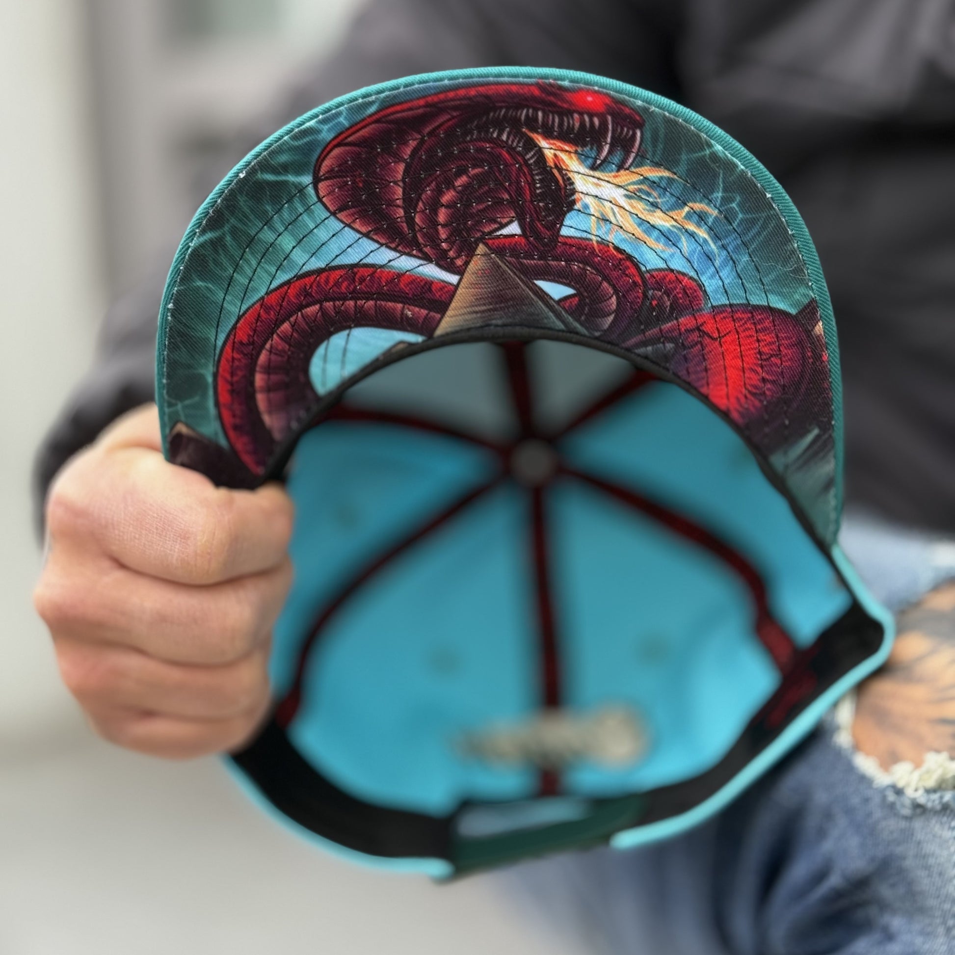 Man holding the Fire Snake snapback and shows the camera the bottom design with a red snake visible from the headwear brand Capiche