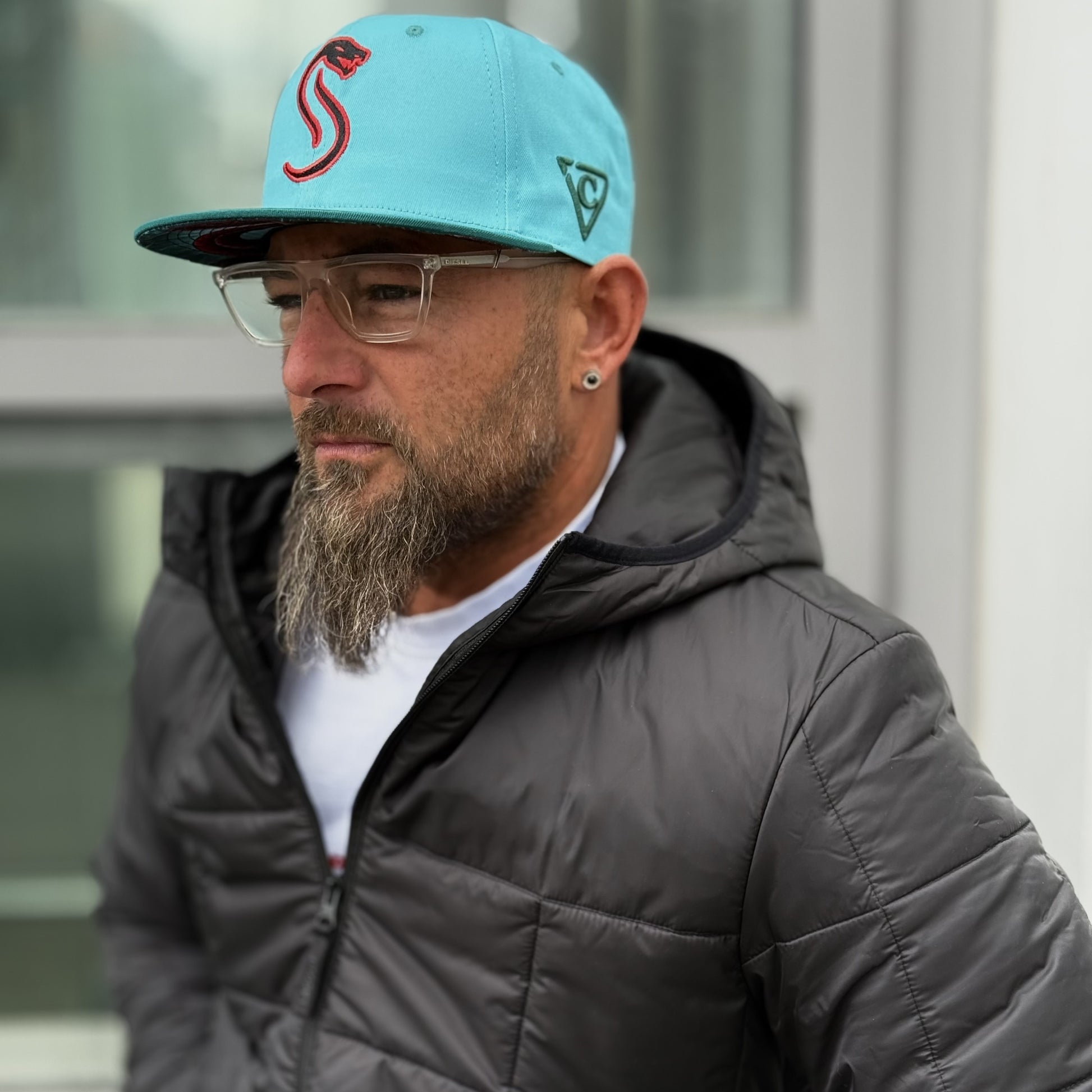 Bearded man standing outside with a jacket on and a blue snapback hat on from the headwear company Capiche