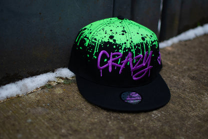 A black snapback with green graffiti spray at the top photographed outside in an urban setting. The snapback is from the brand Capiche Caps