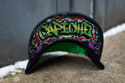 A black snapback with green graffiti spray at the top photographed outside in an urban setting. The snapback is from the brand Capiche Caps