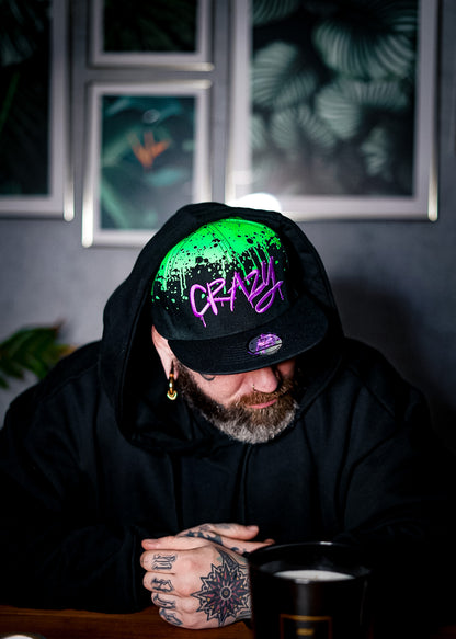 Man with tattoos sitting and wearing a Capiche snapback hat. The snapback hat is black with green graffiti details