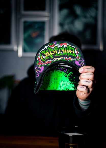 Man holding a snapback to show the bottom design that has the word Capiche on it. The snapback is green and black and is from the headwear company Capiche