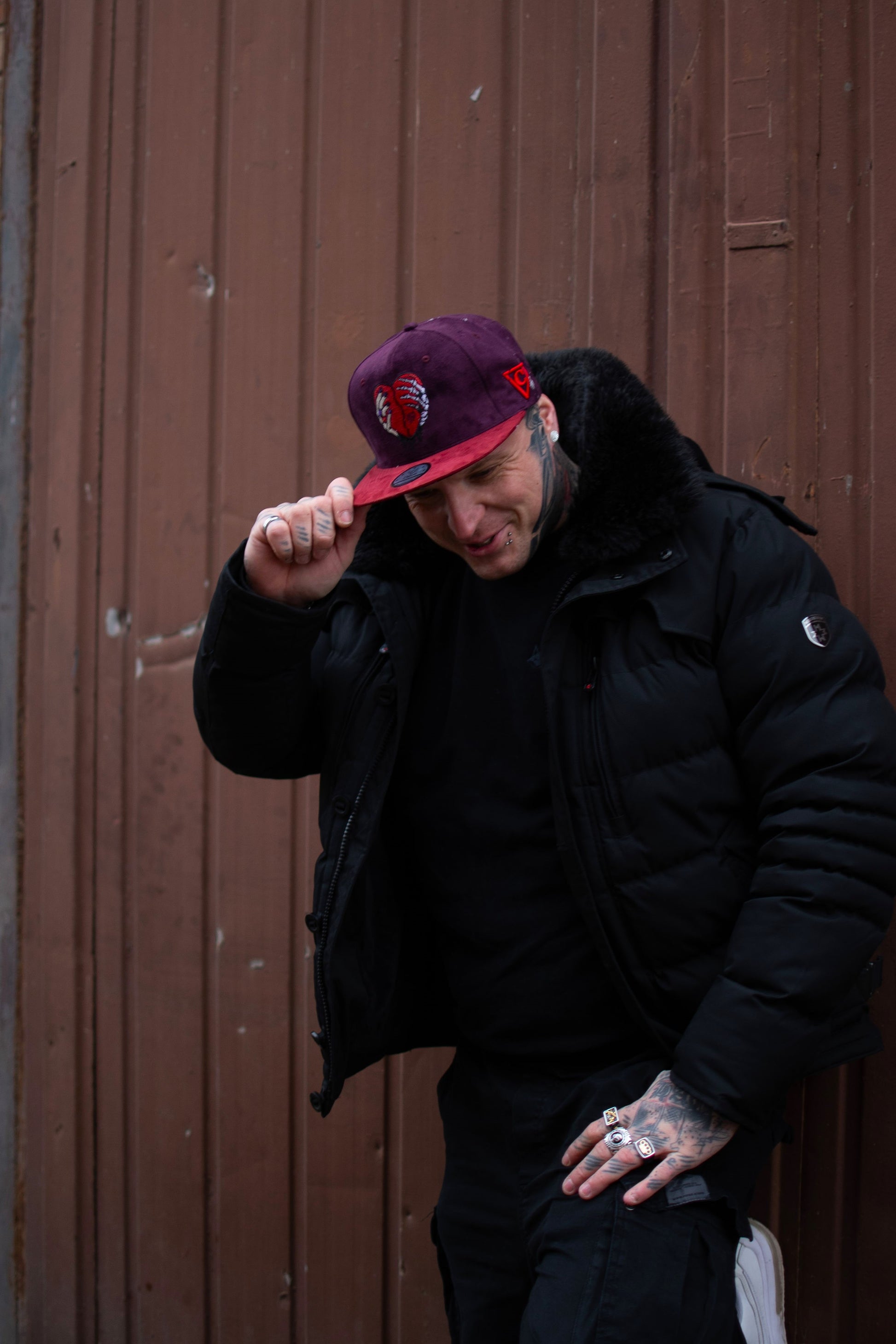 A man standing outside in an urban setting smiling while being photographed with a purple/red snapback on. The snapback is from the headwear company Capiche