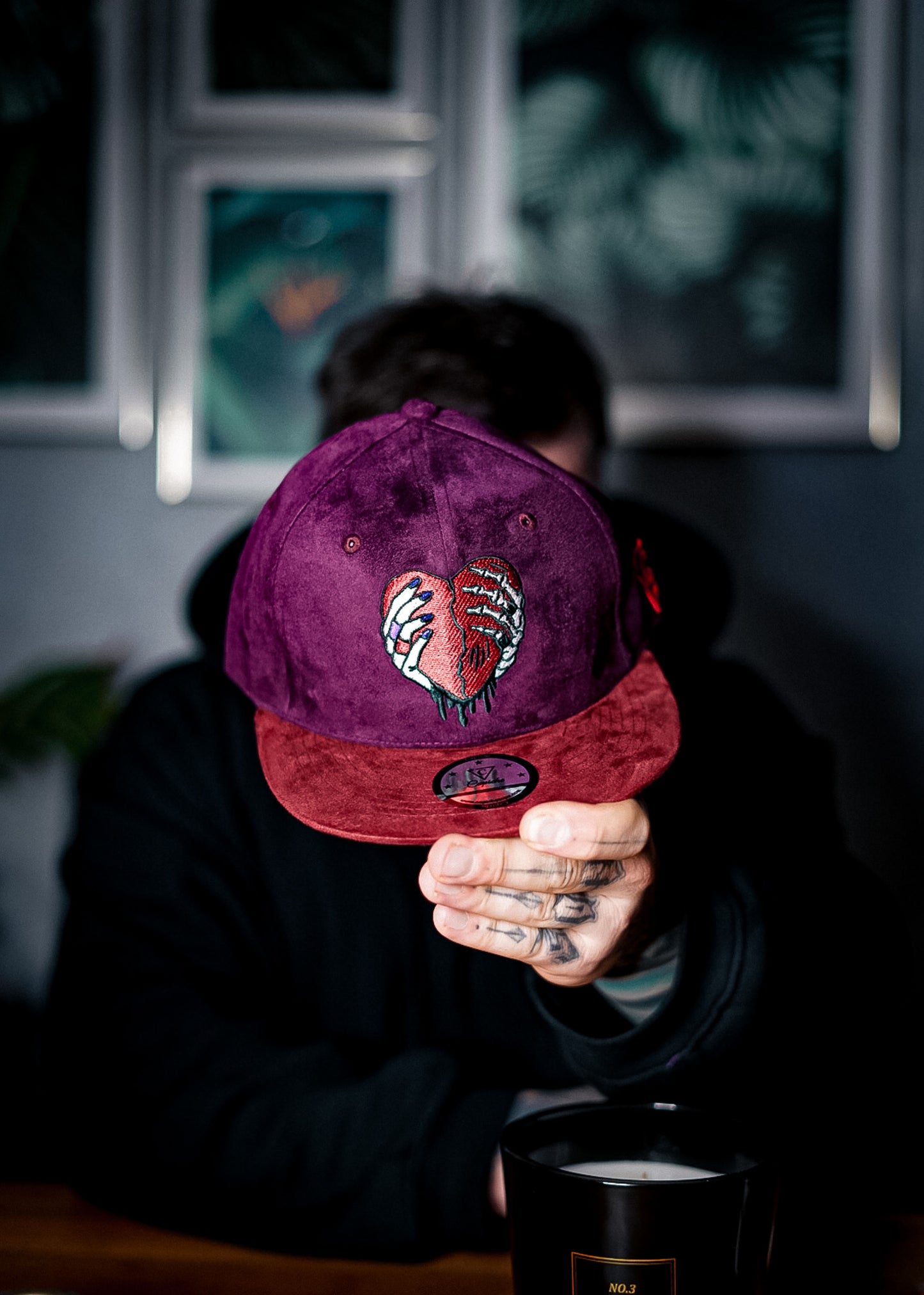 Tattoo model showing the camera the Savior snapback from close up, so the suede fabric is more visible. The front logo of the Capiche snapback is a heart that is being held by a normal hand and a skeleton hand