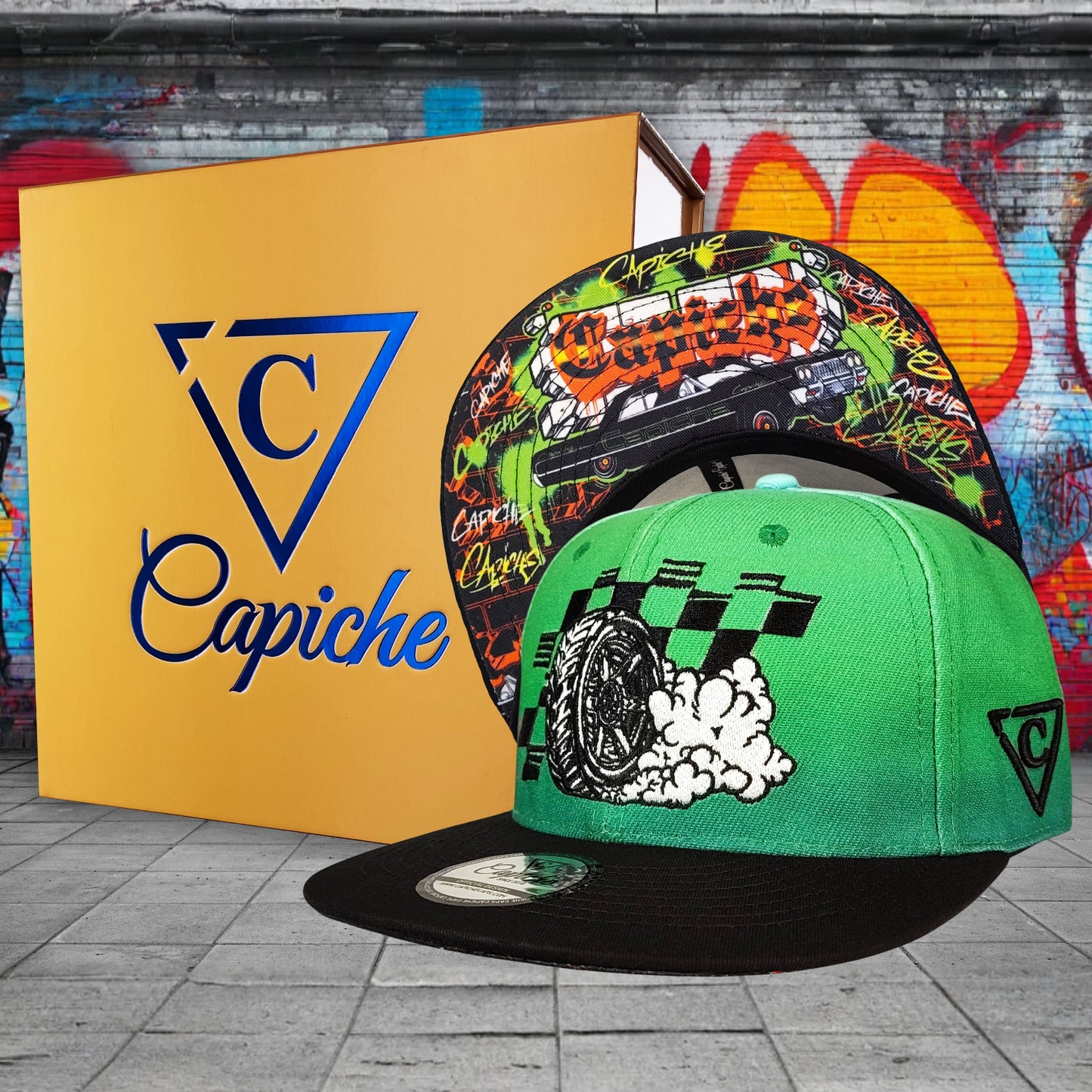 Two caps with colorful designs in front of a Capiche branded box against a graffiti wall.